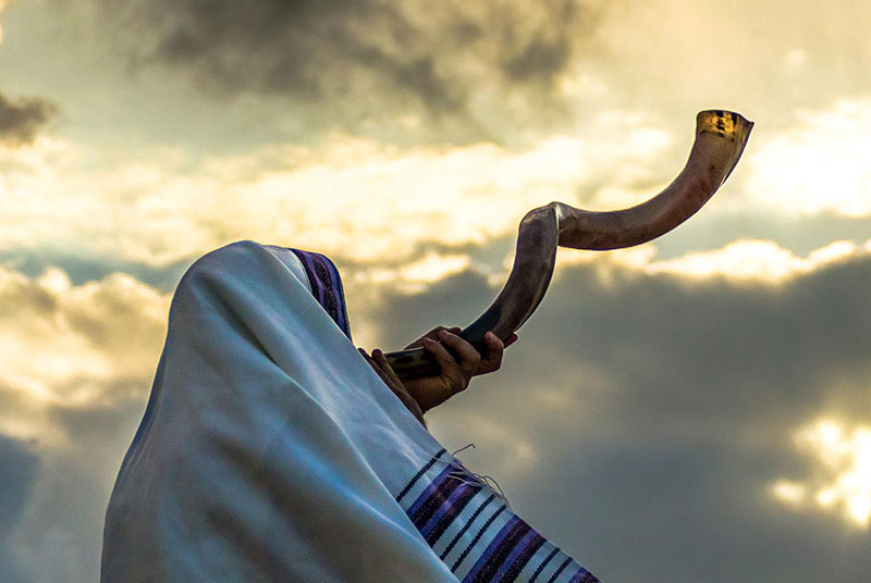 Person in tallit blowing shofar in front of a dramatic, cloudy sky