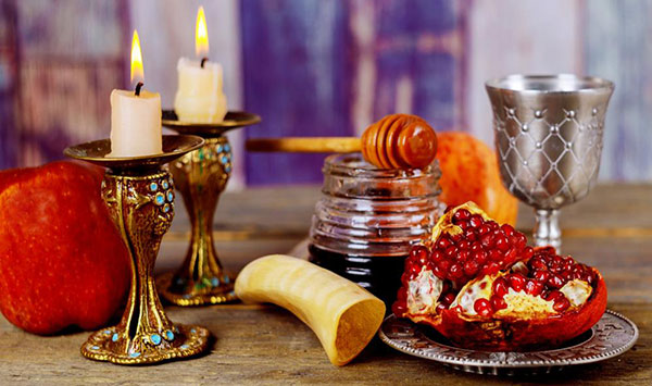 Lit candles on a table with a kiddish cup, apples, honey, a shofar and a new fruit